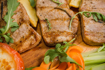 Three pork steaks with fried potatoes and vegetables on a wooden background