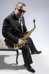 Naklejka premium Portrait of Mature Relaxed Caucasian Saxophone Player in Sunglasses Posing with Saxophone While Sitting on Chair in Studio Environment.