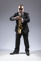 Naklejka premium Music and Musicians Ideas and Concepts. Full Length Portrait of Caucasian Mature Saxophone Player in Sunglasses Playing the Instrument Against White.