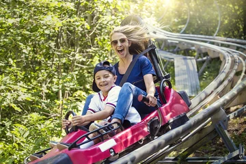 Poster Amusementspark Glimlachende vrouwen en haar jongen rijden samen bergafwaarts op een buitenachtbaan op een warme zomerdag. Ze heeft een leuke uitdrukking terwijl ze genieten van een spannende rit op een rode attractie in een pretpark
