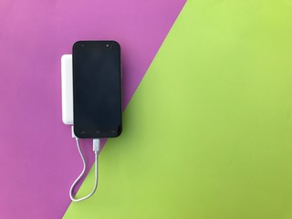 Closeup A black smartphone charging with a white powerbank set on purple and green background.Gadgets on colorful background.Top view with Copy space. Selective focus.