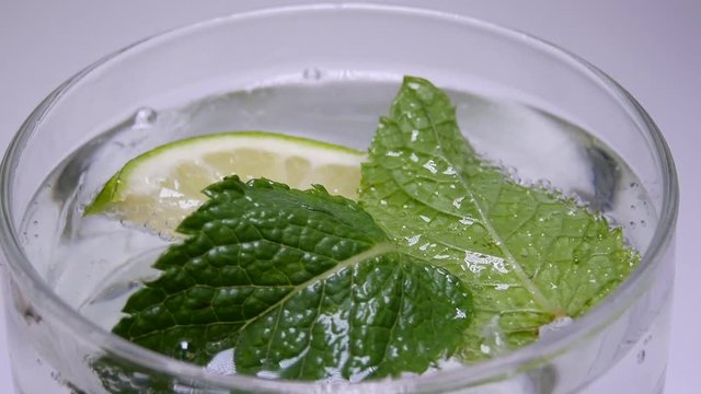 Leaves of Mint and Slices of Lime In Pouring Soda. Soda Pop In Glass. Releasing Bubbles And Foaming In Parkling Water. Aerated Powder Water In Glass On Background of White Wall In Isolated