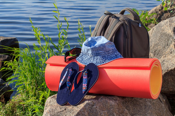 Hiking backpack, hat, flip-flops, sunglasses and camping mat on rock with sea shore background