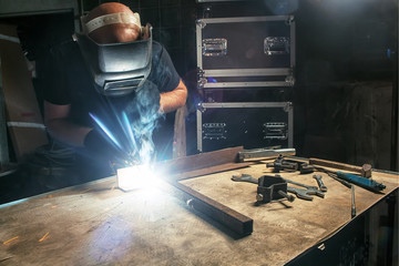 A bald, strong man in a black T-shirt creates with his own hands a mettalical product and welds a...