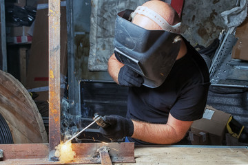 A bald, strong man in black work clothes is welding a metal arc welding machine in a warehouse, against a backdrop of equipment boxes, tools