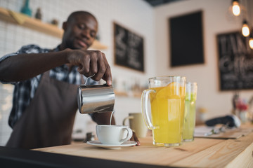 handsome african american barista pouring milk into coffee on bar counter in cafe
