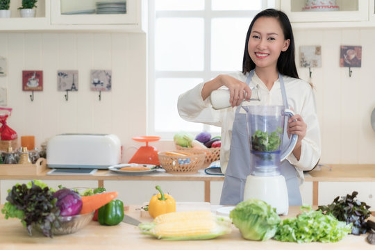 Asian woman making green smoothies with blender home in kitchen. Healthy raw eating lifestyle concept portrait of beautiful young woman preparing drink with spinach, carrots, celery etc.