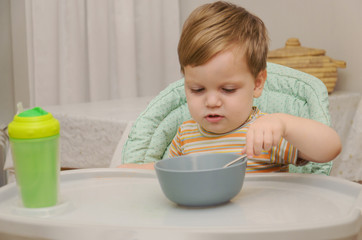 little blond boy in a striped T-shirt eats porridge with a spoon from a gray plate