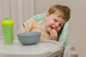 little blond boy in a striped T-shirt does not want to eat a porridge with a spoon from a gray plate.