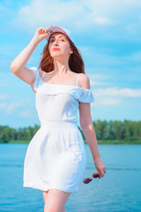 Fototapeta na wymiar A beautiful well-groomed secured young woman in a casual style of clothes, a knitted cape, a white dress and a cap rests by the water for the weekend and feeling freedom