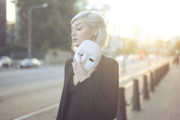 Young blond woman taking off a mask. Pretending to be someone else concept. outdoors on sunset.