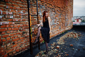 Red haired punk girl wear on black dress at the roof against brick wall with iron ladder.