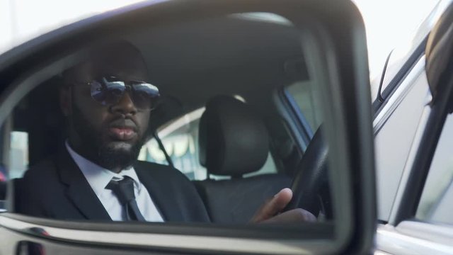 Afro-American in sunglasses driving car, looking in side-view mirror, bodyguard