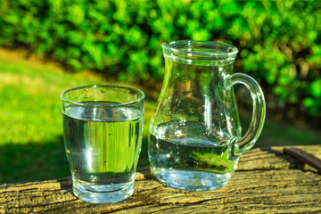 Glass and pitcher with pure water on wooden log, green grass, trees in the background, bright sunny day, healthy diet, hydration, cleansing