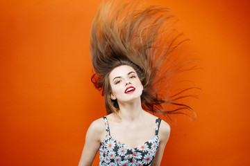 Temperamental Woman with Long Dark Hair Wearing Dress with Flowers is Having Fun in Studio on Orange Background.