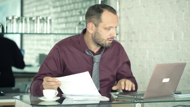 Upset, unhappy businessman working with documents and laptop by table in kitchen
