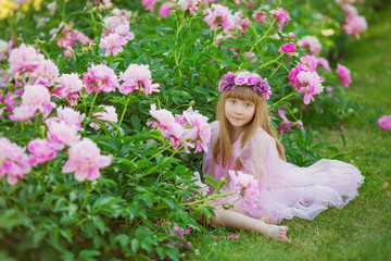 Pretty little girl in a pink dress sits near a pink peonies in a garden