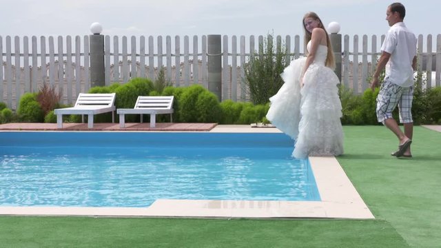 Man pushes young bride from poolside into water as a funny joke. Girl in white wedding dress falls into the pool while trying to pose for camera. Bride falling with splashes in slow motion.