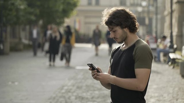 Handsome man waiting in a public urban place for friends texting on social media app on his smartphone