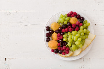 Top view on assortment of juicy fruits on white plate and white table background. Organic raspberries, apricots, melon, cherries - summer dessert or snack. healthy eating concept.