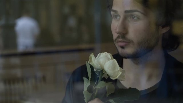 Anxious young man in love waiting for girlfriend with flowers sitting by a window in a pub