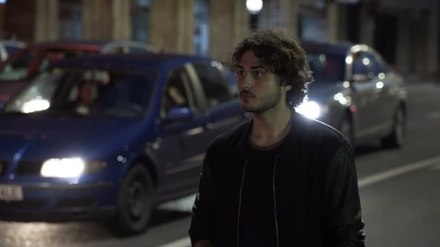 Teenager Man Waiting And Checking Smartphone In The Night For Uber Car Order On Urban Street With Car Traffic