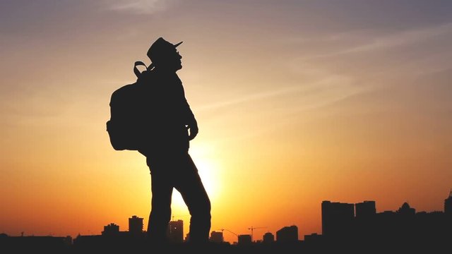 Traveler silhouette with a backpack and cowboy hat standing on the top of hill. City in the background. The sun sets over the horizon. A man looks ahead, raise his arms up. Travel, holidays, advanture