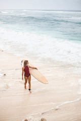 Fototapeta na wymiar Beautiful surf girl in dark-red swimming suit is walking on the beach with a surfboard and looking away, located on the left side of the frame