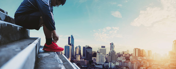 Asian Guy Tying Running Shoe, Preparing for Running, with Panoramic City background in Sunrise