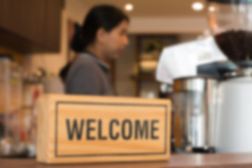 Blurred background of female barista are preparing coffee drink behind a welcome wooden sign in the cafe or coffee shop.