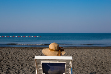 A young girl on the beach, enjoying a sun break by the sea on a sidewalk, a hat on her head, reading a book, in the background a blue sea