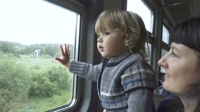 Train departs from station. Little curious boy and her mother looking out of window in train. Drops of rain on glass. It's raining outside, people reflecting in glass.
