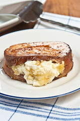 Baked, organic, apple, cinnamon stuffed french toast with powdered sugar on a round, white plate with a blue rim and an artisan serving utensil all on a white table cloth with blue stripes