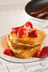 Baked, stuffed french toast, local, ripe strawberries, strawberry butter on the stack on a round, white plate with a blue rim, a pan of cooked bacon, on a blue striped white cloth 