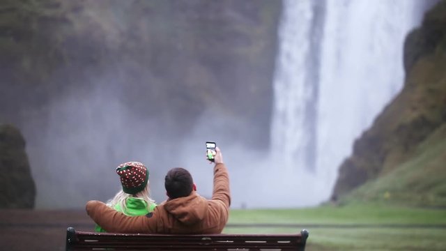 Back view of young couple sitting on the bench and taking selfie photo on smartphone. Skogafoss waterfall in Iceland.