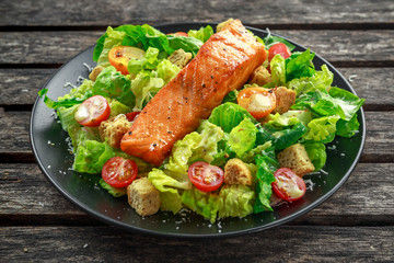 Caesar Salmon Salad with croutons, parmesan cheese, tomatoes, dressing and pepper in a black plate on wooden table.