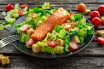 Caesar Salmon Salad with croutons, parmesan cheese, tomatoes, dressing and pepper in a black plate on wooden table.