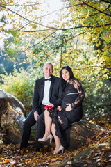 A young pregnant cheerful woman sits on a bench in the park near her happy husband who keeps little red shoes