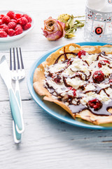 Homemade pancakes or Russian pancakes with chocolate sauce, whipped cream and raspberries on a plate on a white wooden background.
