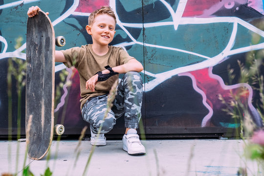 Boy with skateboard sits near graffiti painted wall