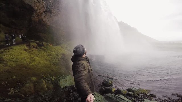 Young tourist man with selfie stick filming on gopro camera powerful waterfall Seljalandsfoss in Iceland in overcast day