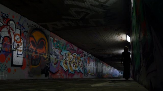 A young woman walking beside an underpass wall. The underground passageway is spray painted all over, and fluorescent light illuminates the girl as she walks towards the camera.