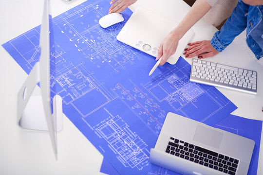 Two young woman standing near desk with instruments, plan and laptop.