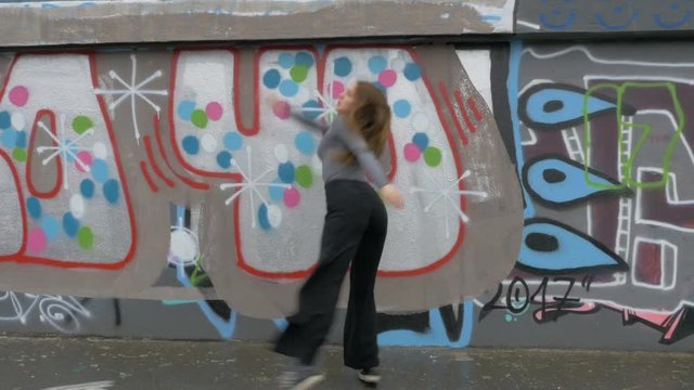 A street dancer dances behind a spray painted city wall as rain falls.