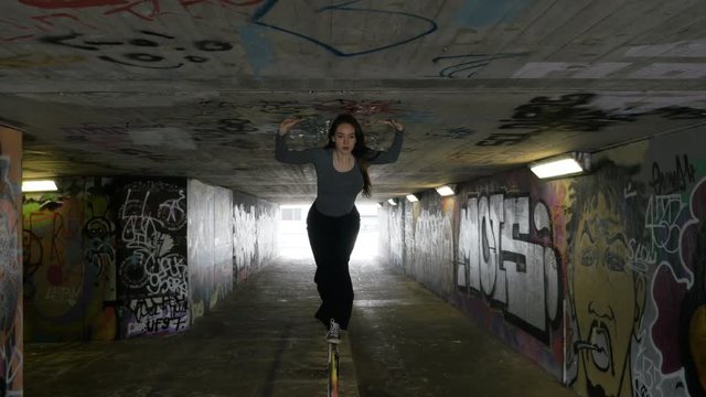 A girl dances and balances herself on a handrail in an urban passageway. The camera moves around her.