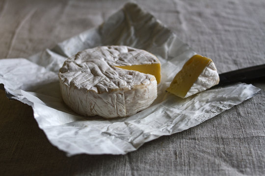 The head of the Swiss Camembert cheese and a triangular piece of cheese on a black knife on foil-wrapped packaging on a baggy fabric background.
