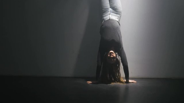 A dancer does the splits from a handstand position. The dance room is smokey and dark.