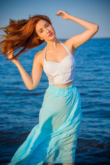 Redhead model wearing tender pastel dress posing on a sea. Young and fashionable  woman walking on the shore. Young girl go to beach in a white tones. Pastel colors. The concept of femininity. 
