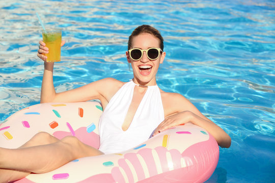 Beautiful young woman with glass of cocktail relaxing on inflatable donut in pool