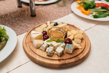 Cheese plate with cherries and honey on round wooden board plate on celebratory dinner table, selective focus. Camembert, DorBlu, Emmental, Brie on cutting board